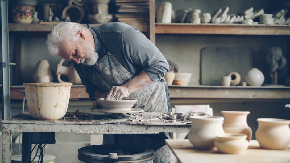 Elderly man skillfully shaping clay on potter's wheel in a pottery workshop filled with artifacts.