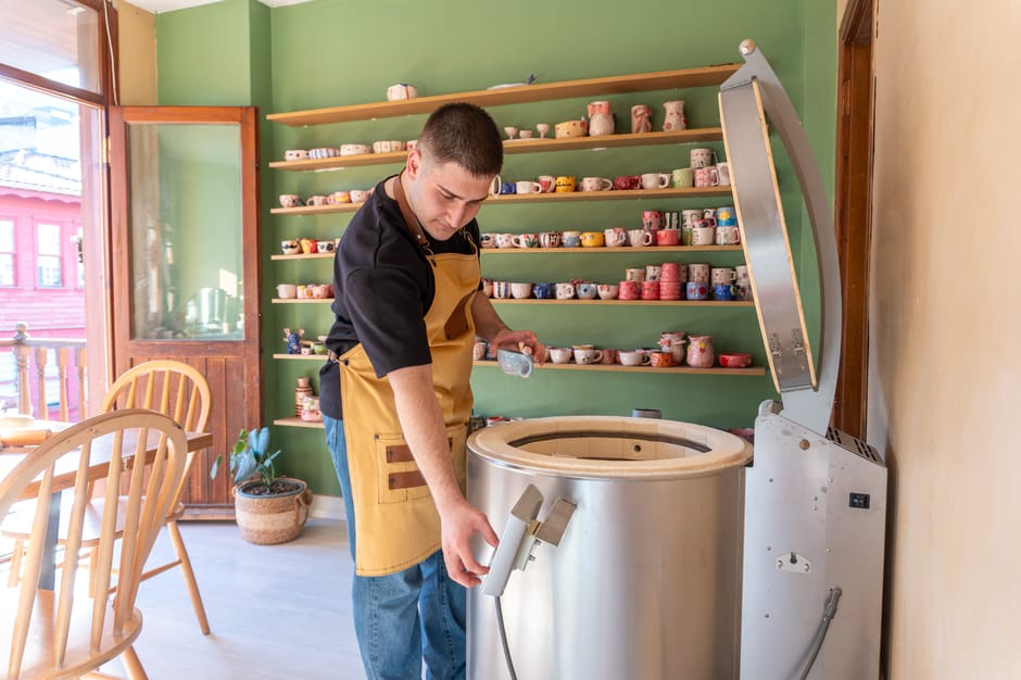 A man working with a kiln in a pottery studio, surrounded by colorful ceramics on display.